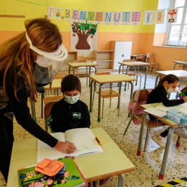 Teacher supports three students sitting in an otherwise empty classroom, all wearing face masks.